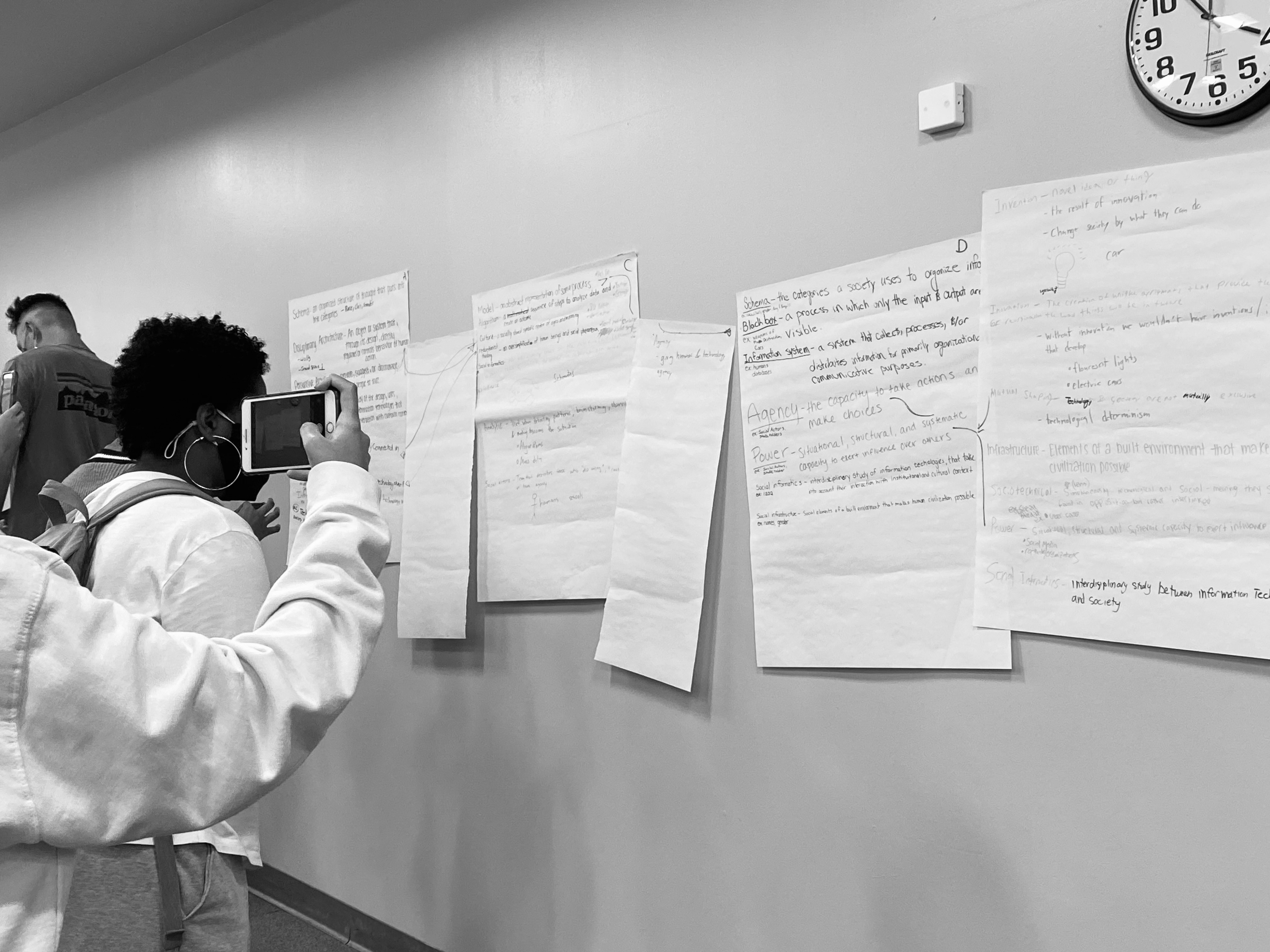 College students looking at large, poster sized pieces of paper stuck on a wall. The pieces of paper have hand written notes over them. A student is using a cell phone to take a picture of the notes.