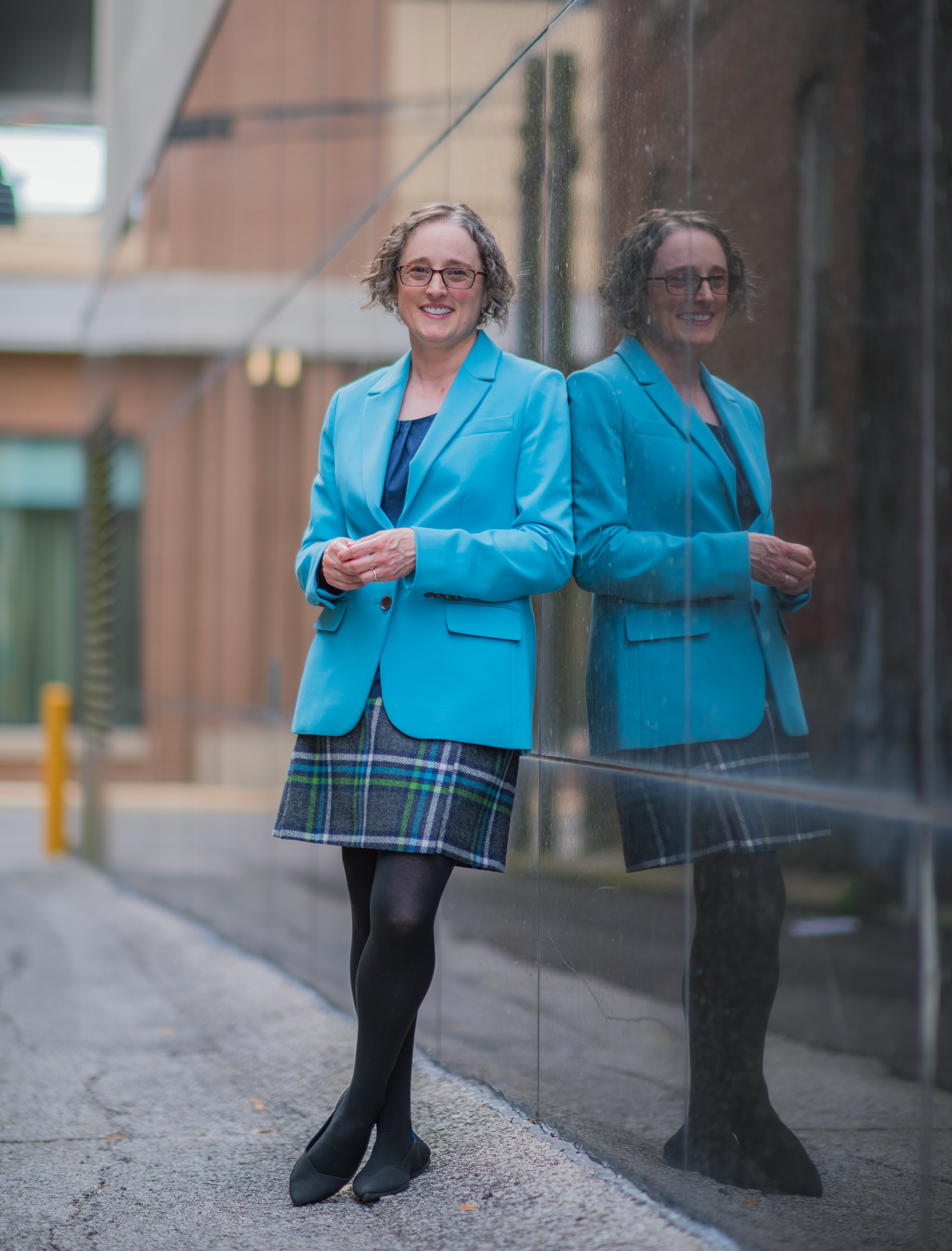Image of a person standing outside in an alley. Person is wearing a blazer and tweed skirt. Person has wavy hair and is wearing glasses. Person is smiling.